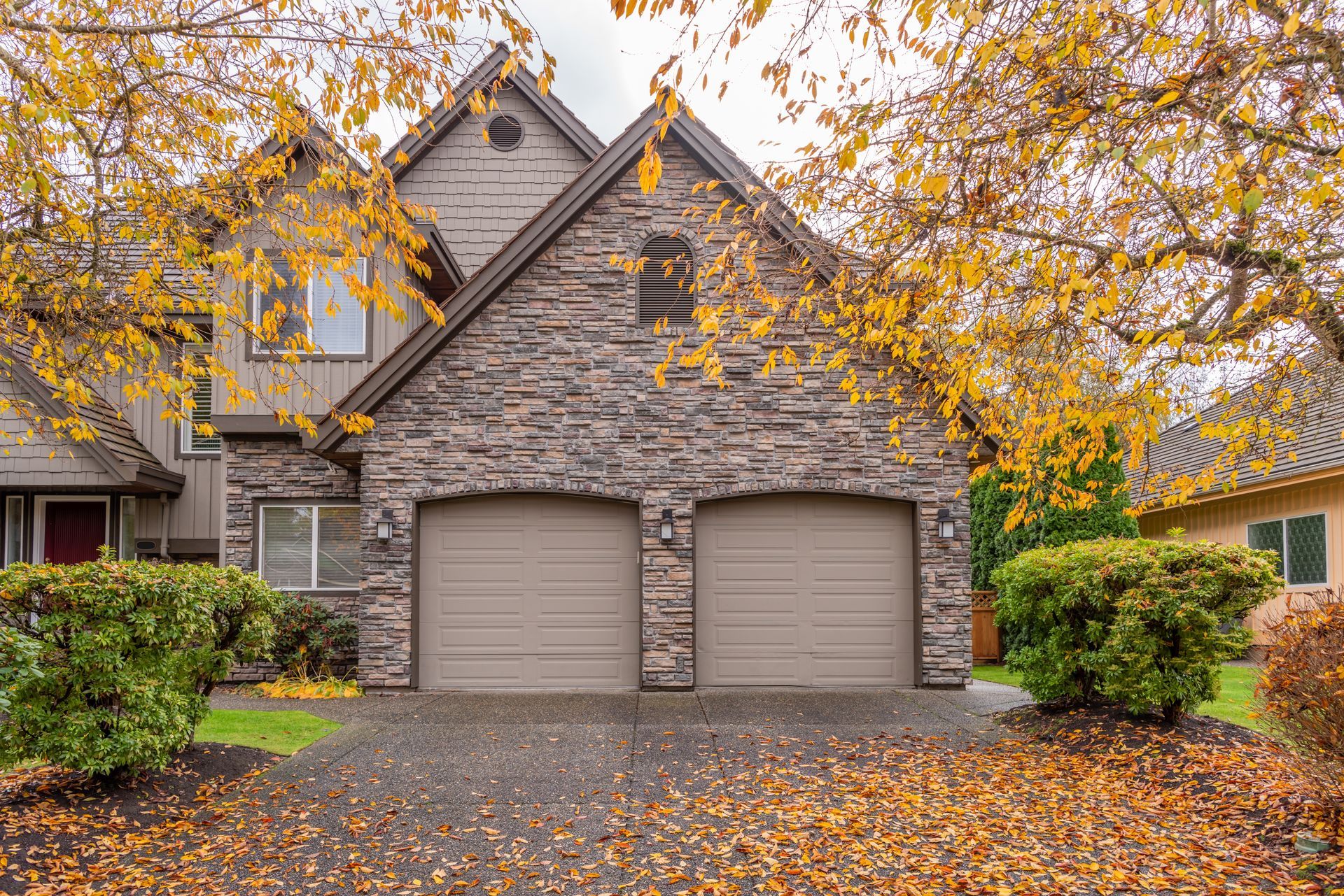 Suburban stone house with twin garage doors, framed by autumn trees and fallen leaves