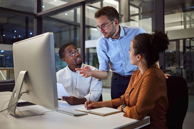 Three coworkers discussing at a desktop computer in a glass-walled office, one pointing at the screen
