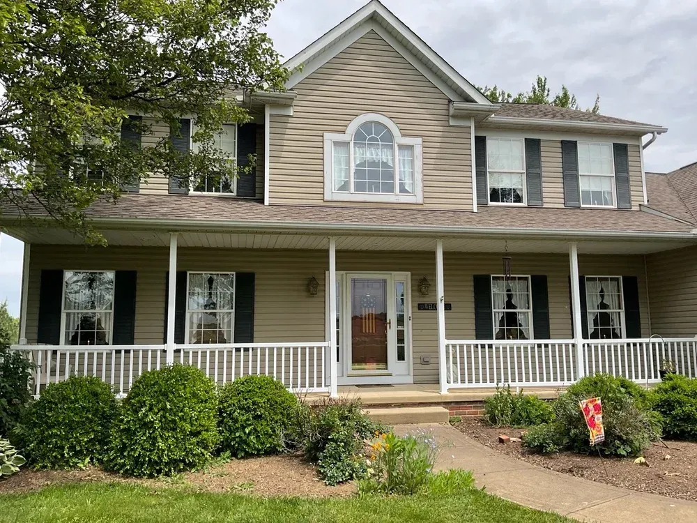 Tan two-story house with a white porch. Black shutters and a brown front door. Landscaping in front.