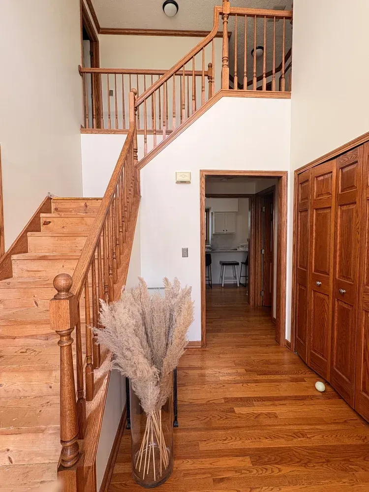 Wooden staircase in a home entryway, leading to an upper level. Hardwood floors, light walls, and a vase with tall pampas grass.