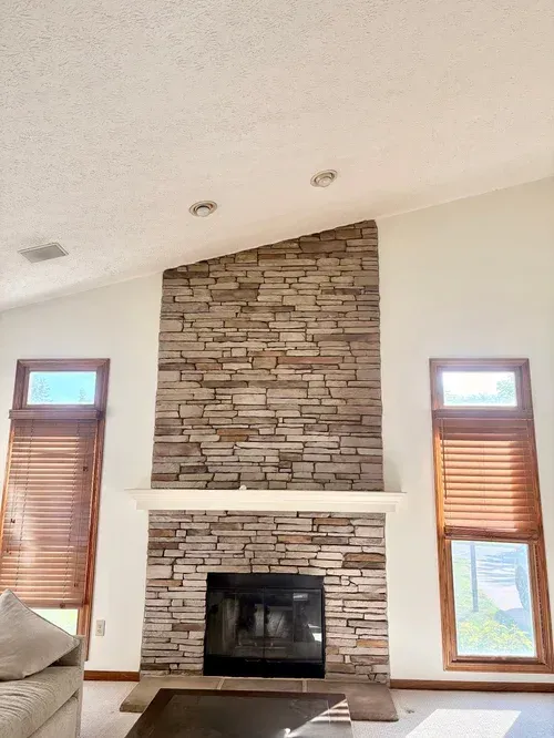Fireplace with stone facade, flanked by windows with wooden blinds, in a white-walled room.