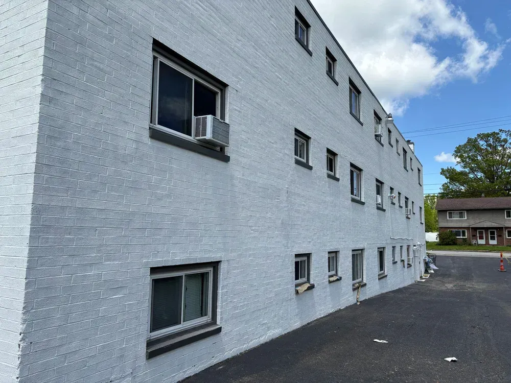 Gray painted brick building with windows and air conditioners, asphalt lot, and blue sky.