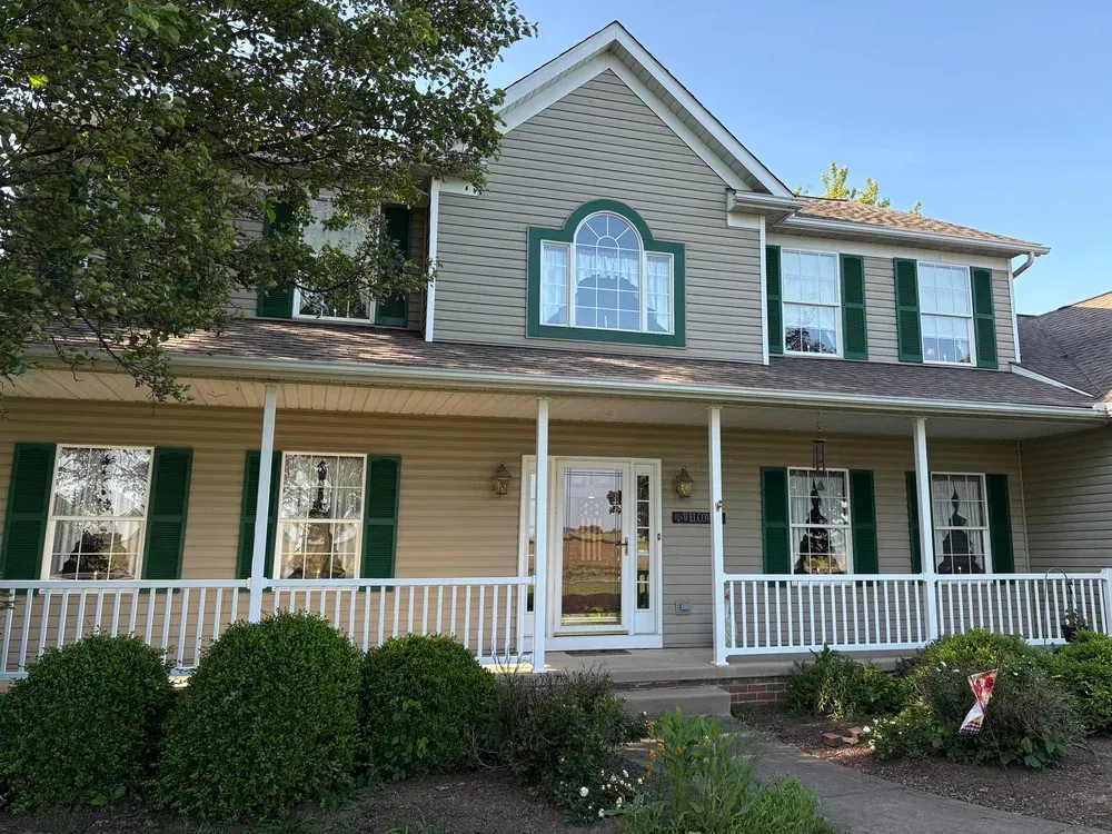 Tan two-story house with green shutters, a porch, and a front yard with bushes.