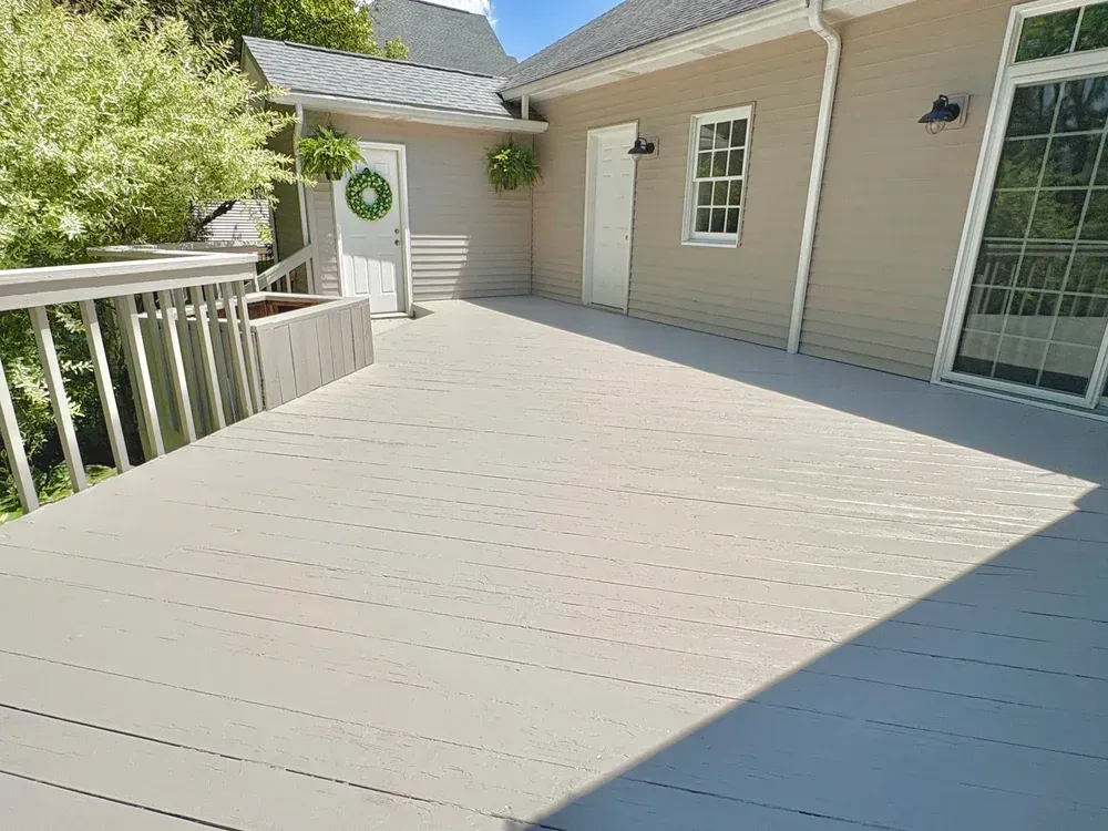 Wooden deck with a light gray surface, connecting to a house with beige siding and a white door.