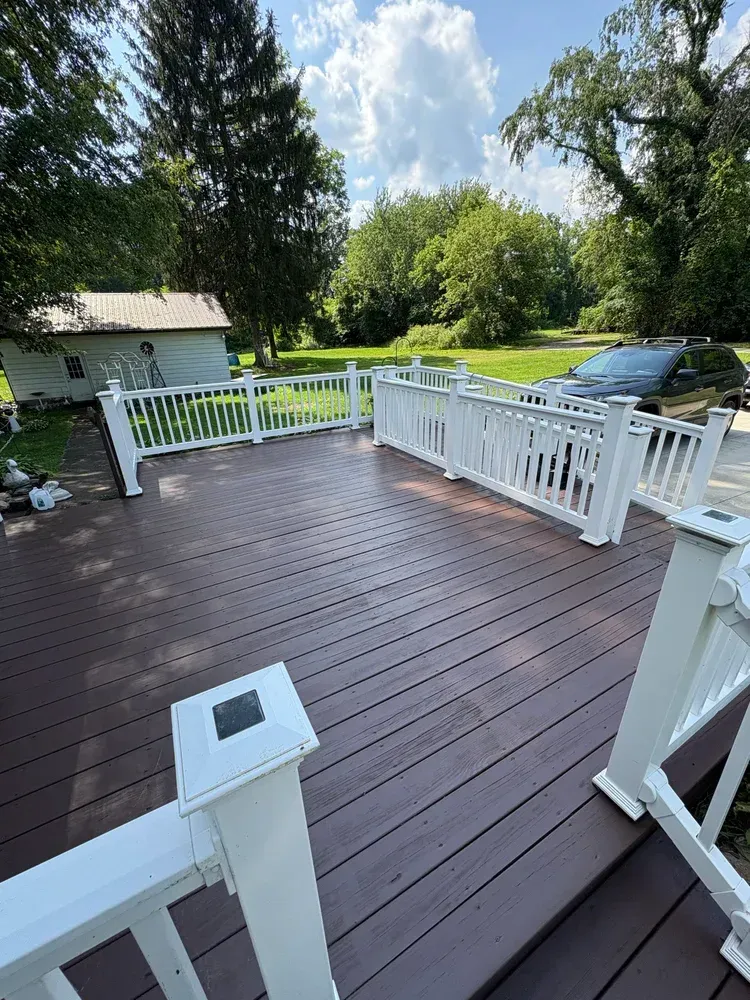 A brown composite deck with white railings and solar post caps. A small house and trees are in the background.