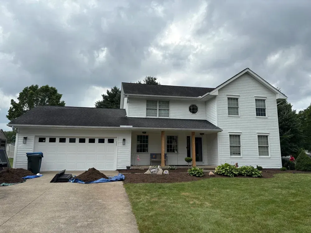 White two-story house with dark roof and garage. Landscaping and overcast sky are visible.