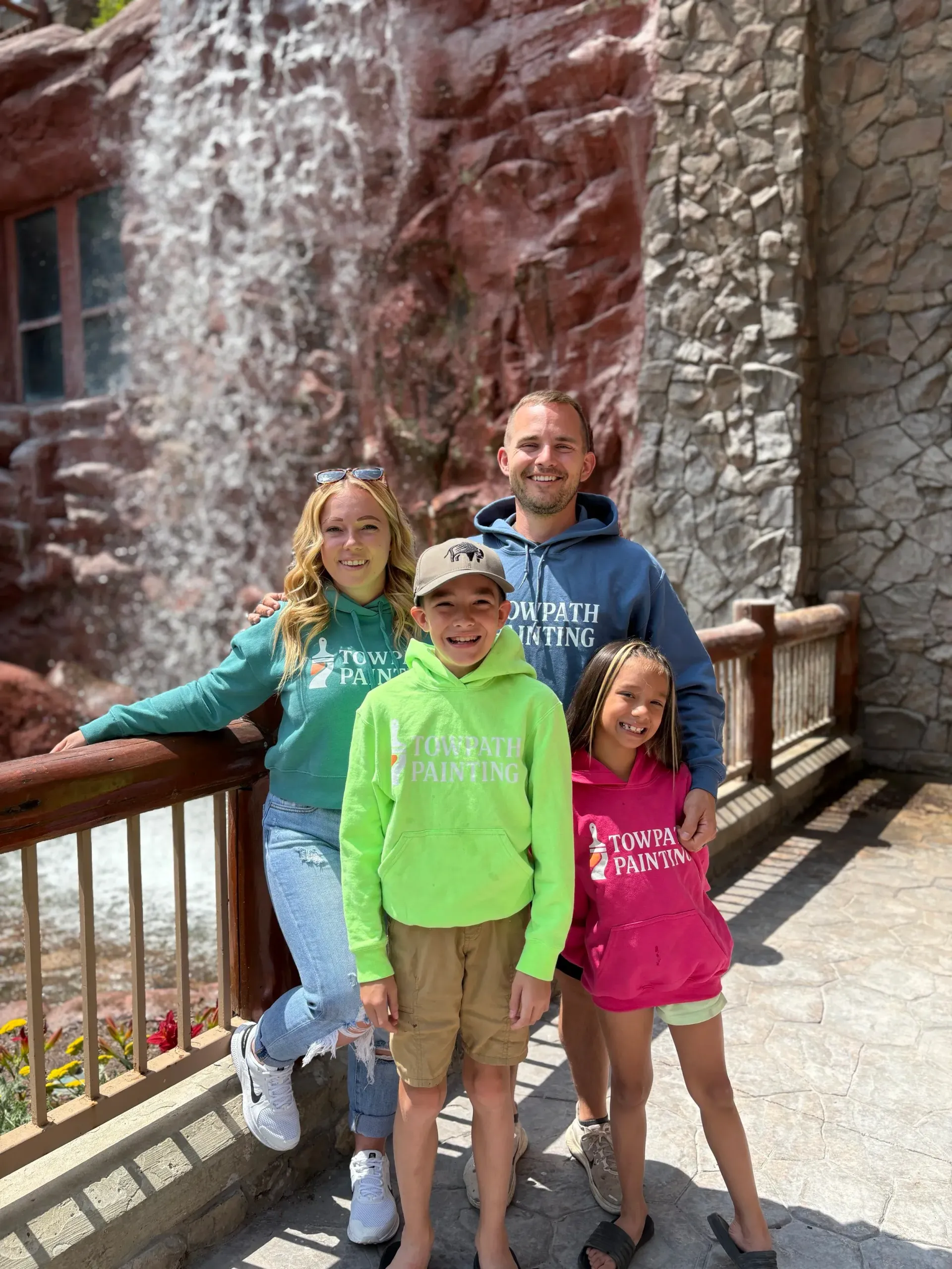 Family posing in front of a waterfall and stone building. They wear matching hoodies; the boy is in green.