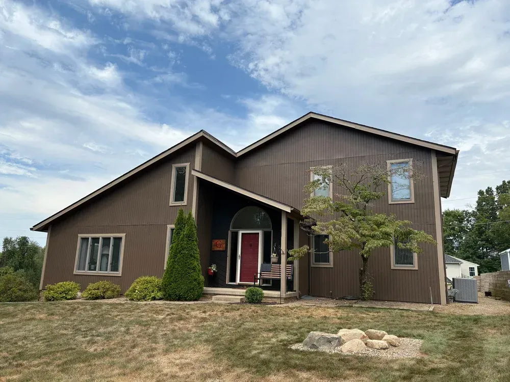 Brown two-story house with red door, windows, and green landscaping under a cloudy blue sky.