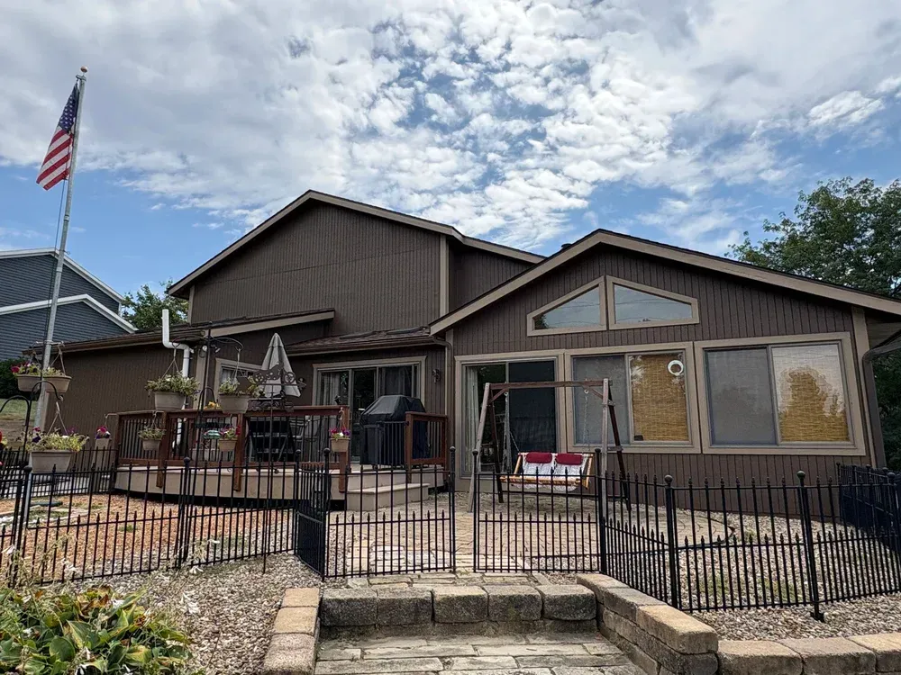 Brown house with deck and flag, patio with a black fence, blue sky.