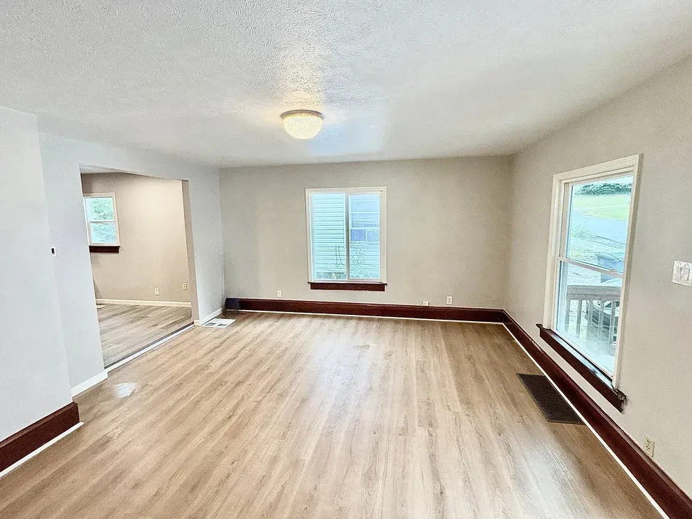Empty room with beige walls, wood-look flooring, and windows with dark wood trim; overhead light.