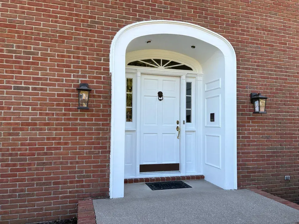 White front door with arched frame, on brick building, two lanterns flank the door.