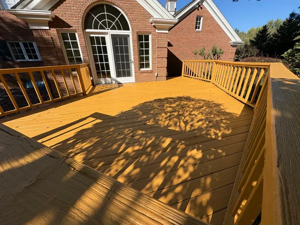 Wooden deck painted yellow, attached to a brick house. Shadows from trees cover the deck.
