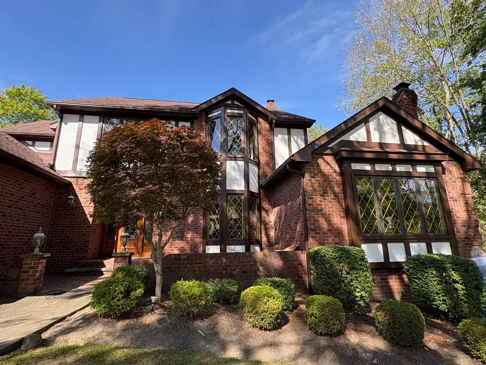 Two-story Tudor style brick home with brown trim and windows. Bushes and small tree in front.