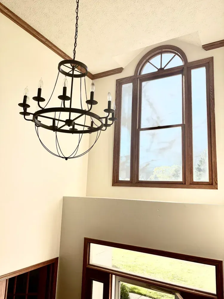 Chandelier hangs near a large window, both in a light-colored room. Brown trim frames the window and ceiling.