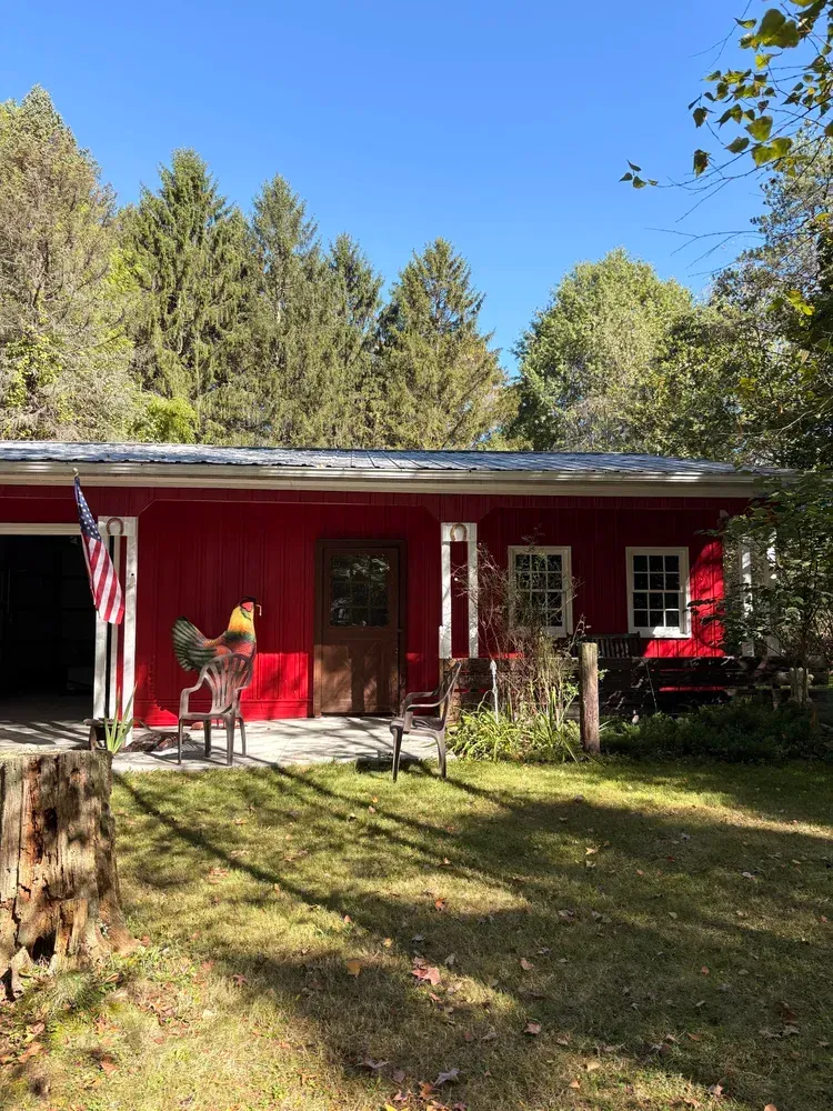 Red barn with an American flag, metal roof, and rooster statue. Sunlight bathes the grassy yard.