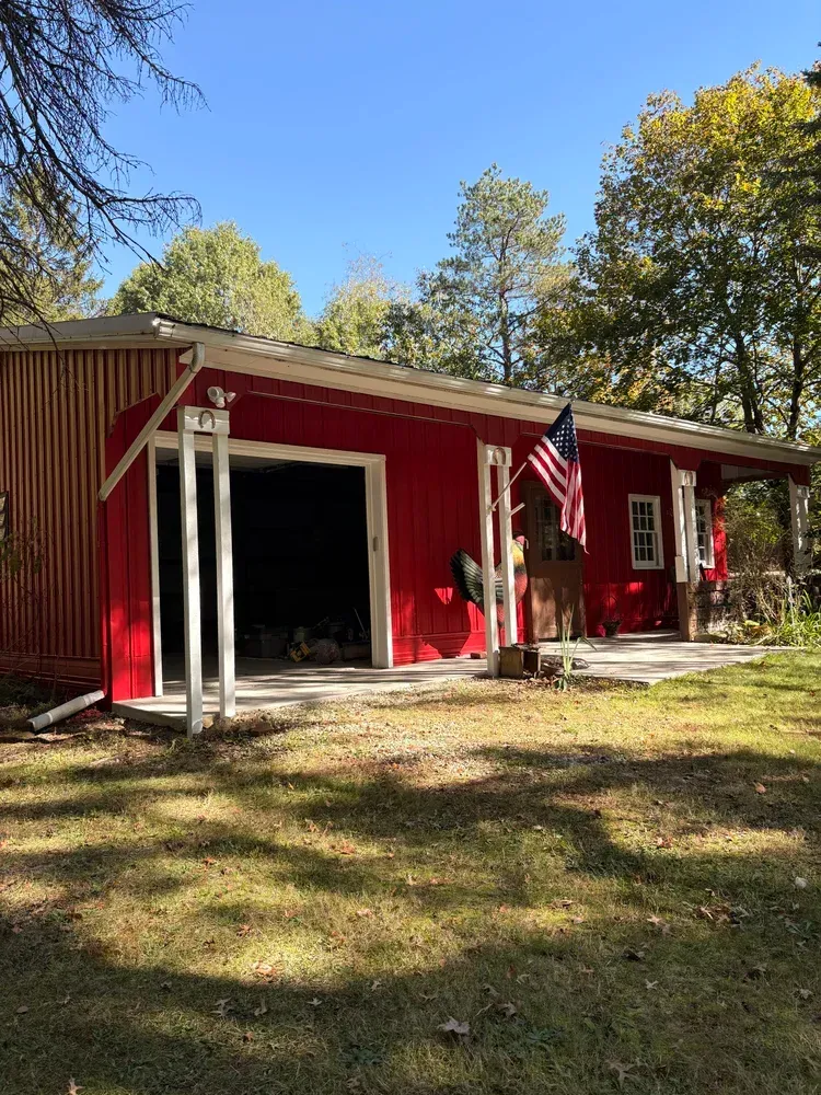 Red building with open garage and porch, American flag, trees, and blue sky.
