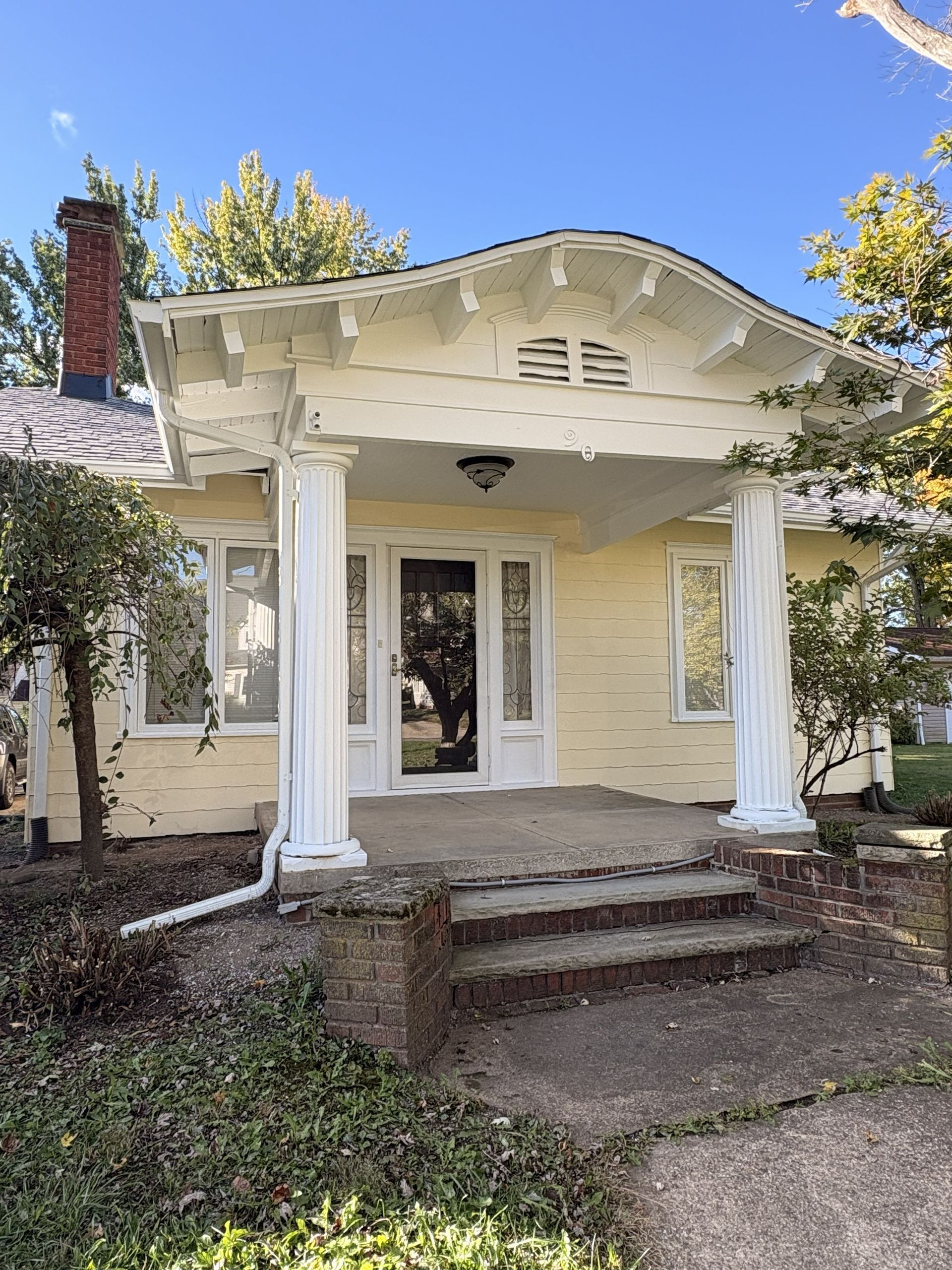 Yellow house with white columns, porch, and steps under a blue sky with trees.