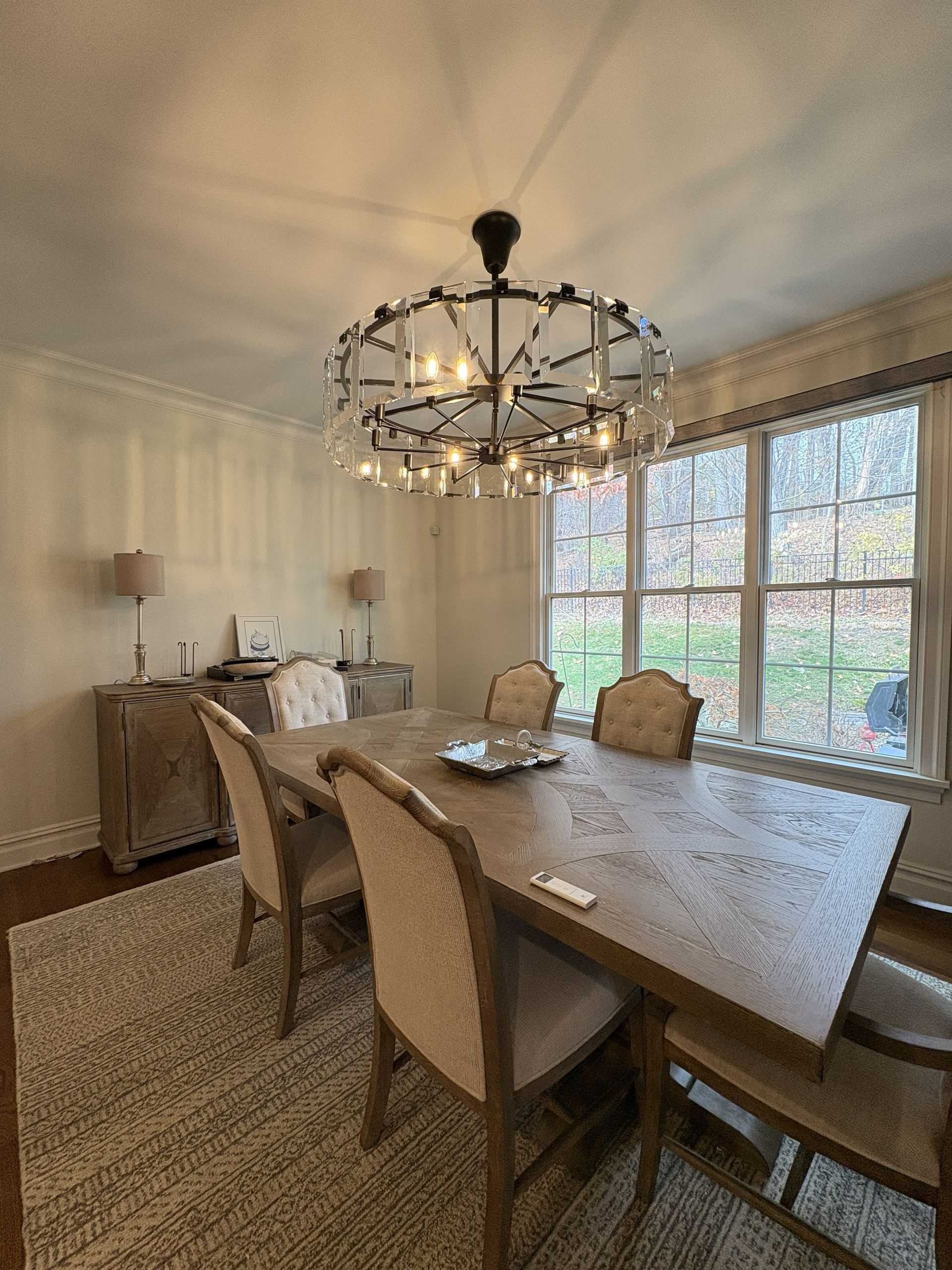 Dining room with wooden table, chairs, rug, and chandelier. Window and sideboards visible.