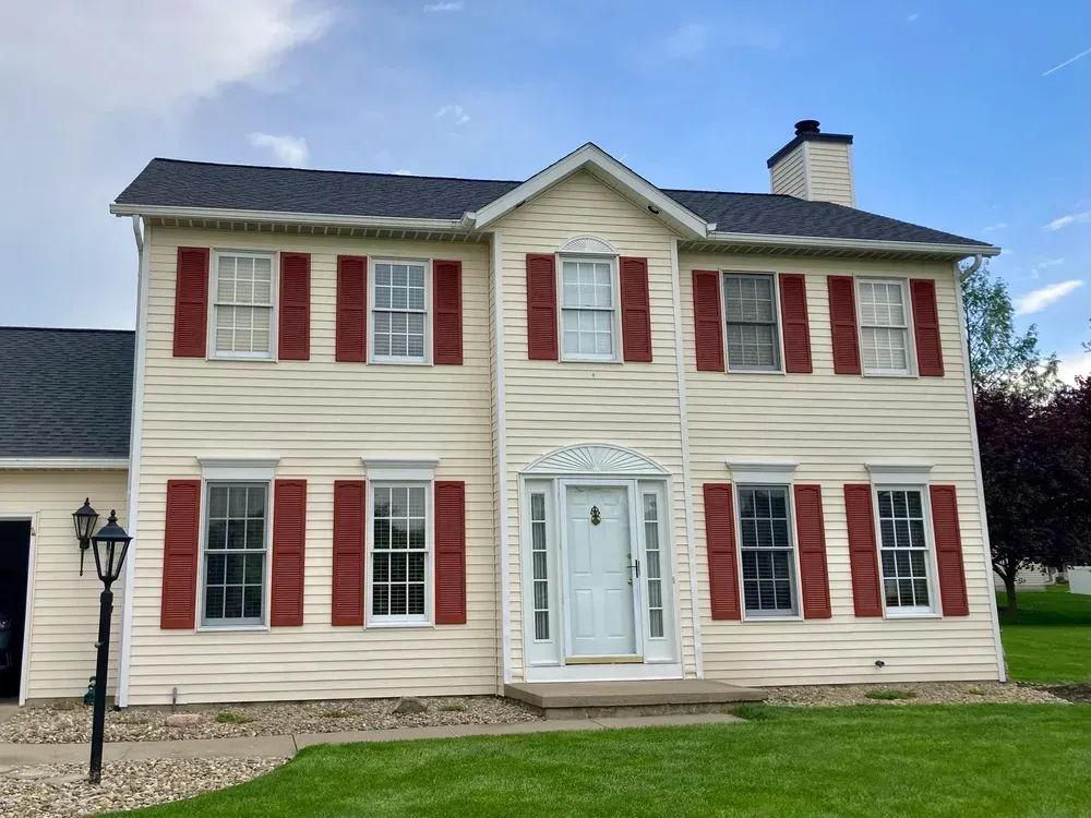 Two-story house with tan siding, red shutters, white trim, and a black roof.