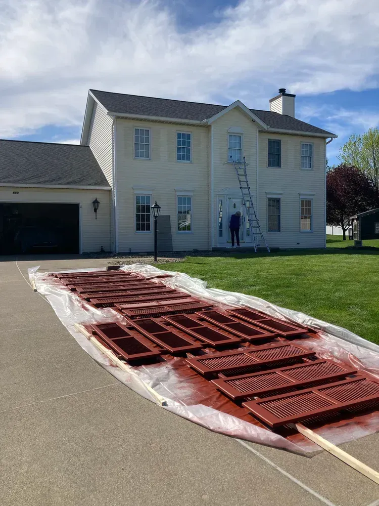 A two-story house with a person on a ladder painting shutters red on a sunny day.