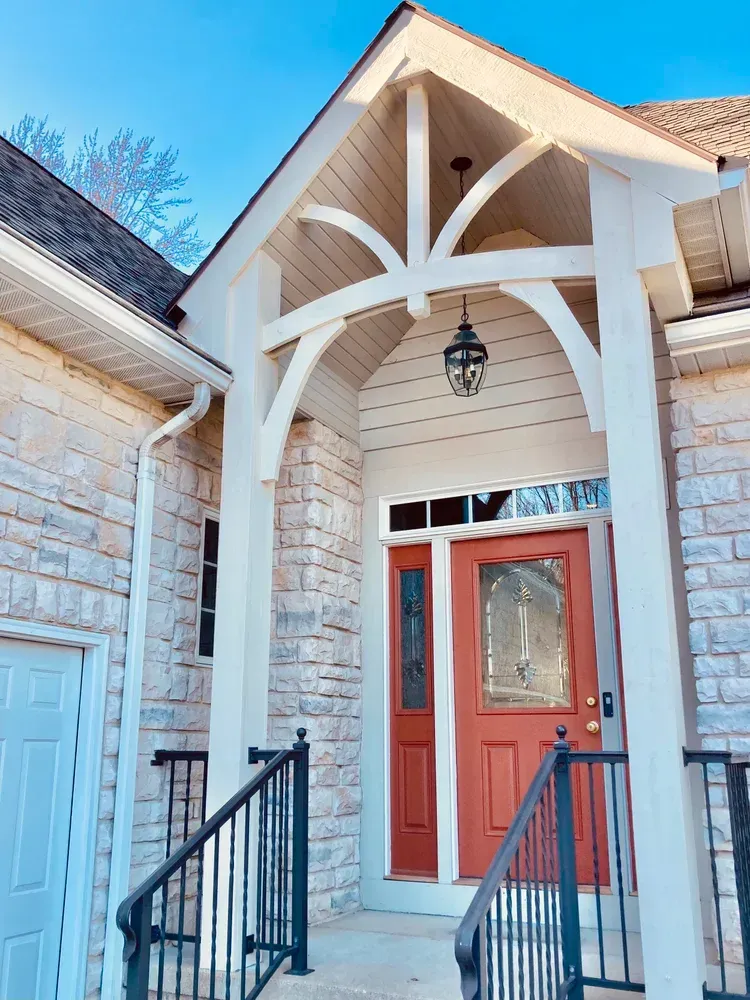 Red door beneath a beige arched porch with black railing and light stone siding.
