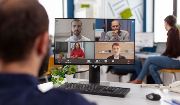 Man in office, facing computer with video call displaying four people; another person sits nearby.