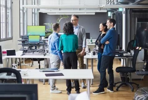 People in an office standing and talking near desks with computers.