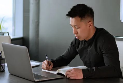 Man in black shirt writing in a notebook, looking at a laptop at a desk.
