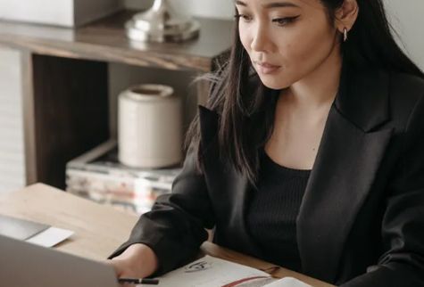 Woman in a black blazer focused on a laptop at a desk with a light wooden top.