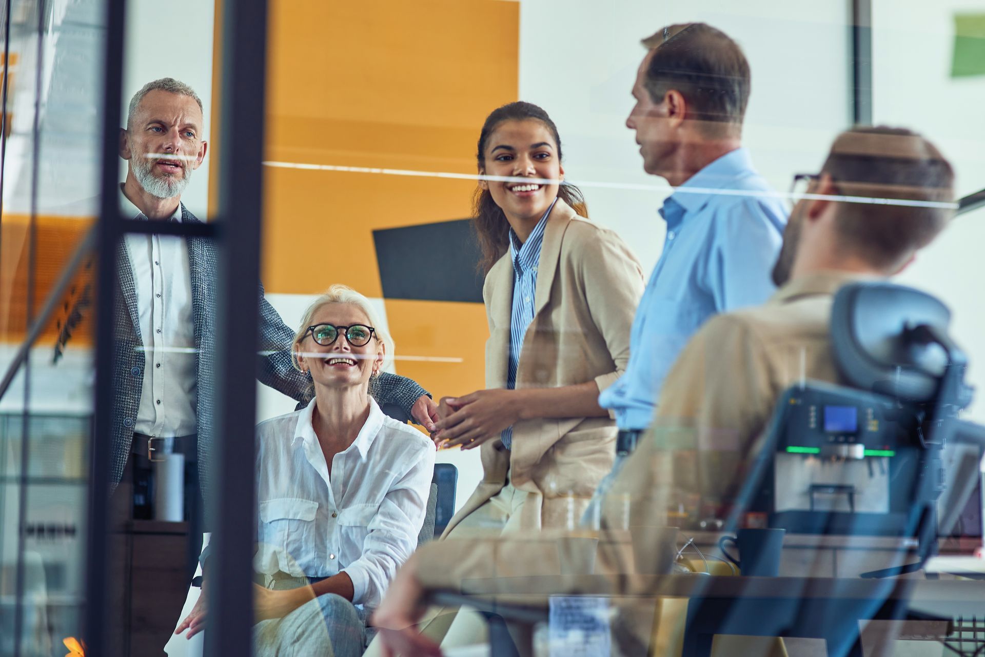 Group of people in an office setting, interacting and smiling. Some are standing, and some are seated.