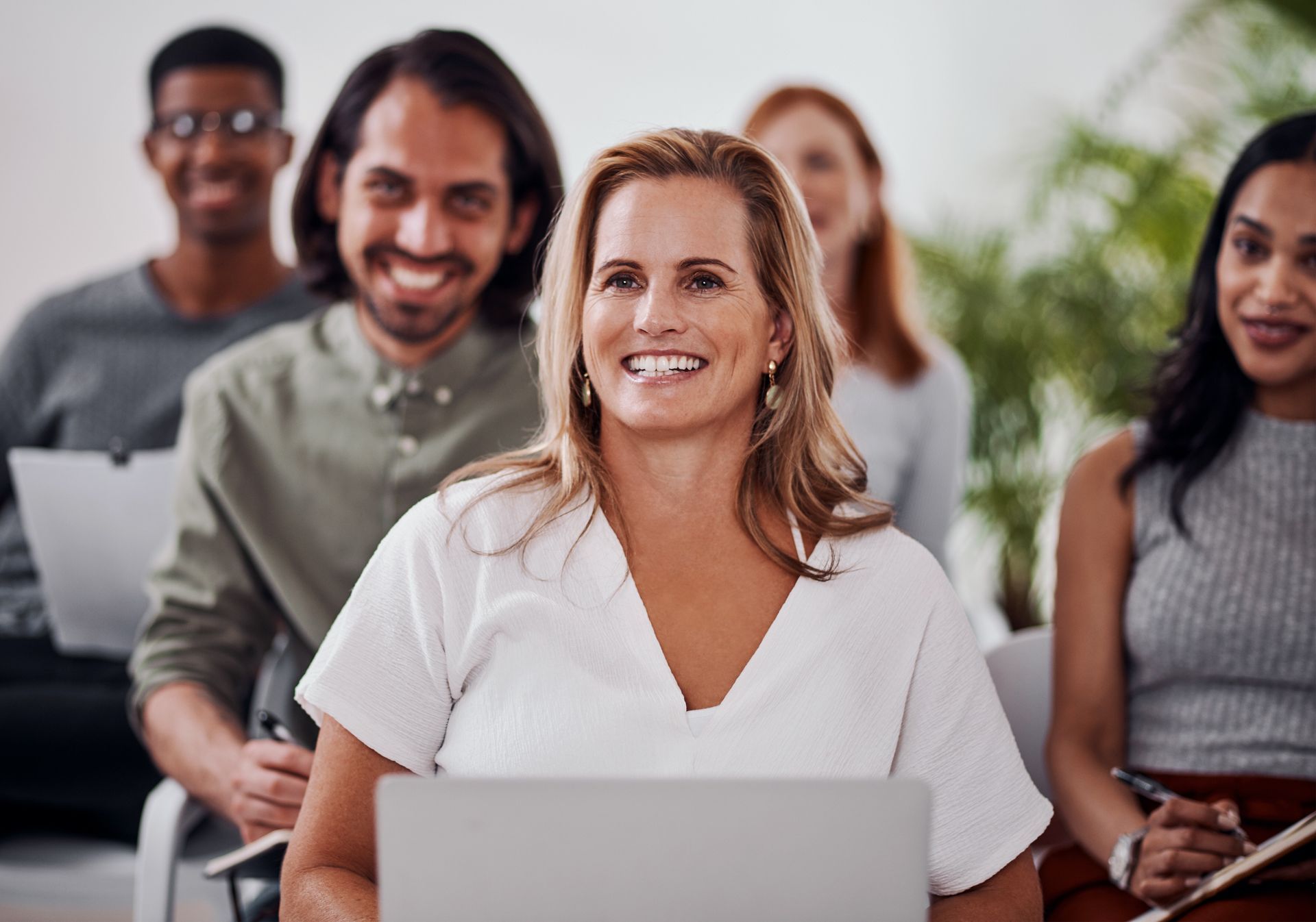 People smiling and listening in a meeting, with laptops and notebooks visible.