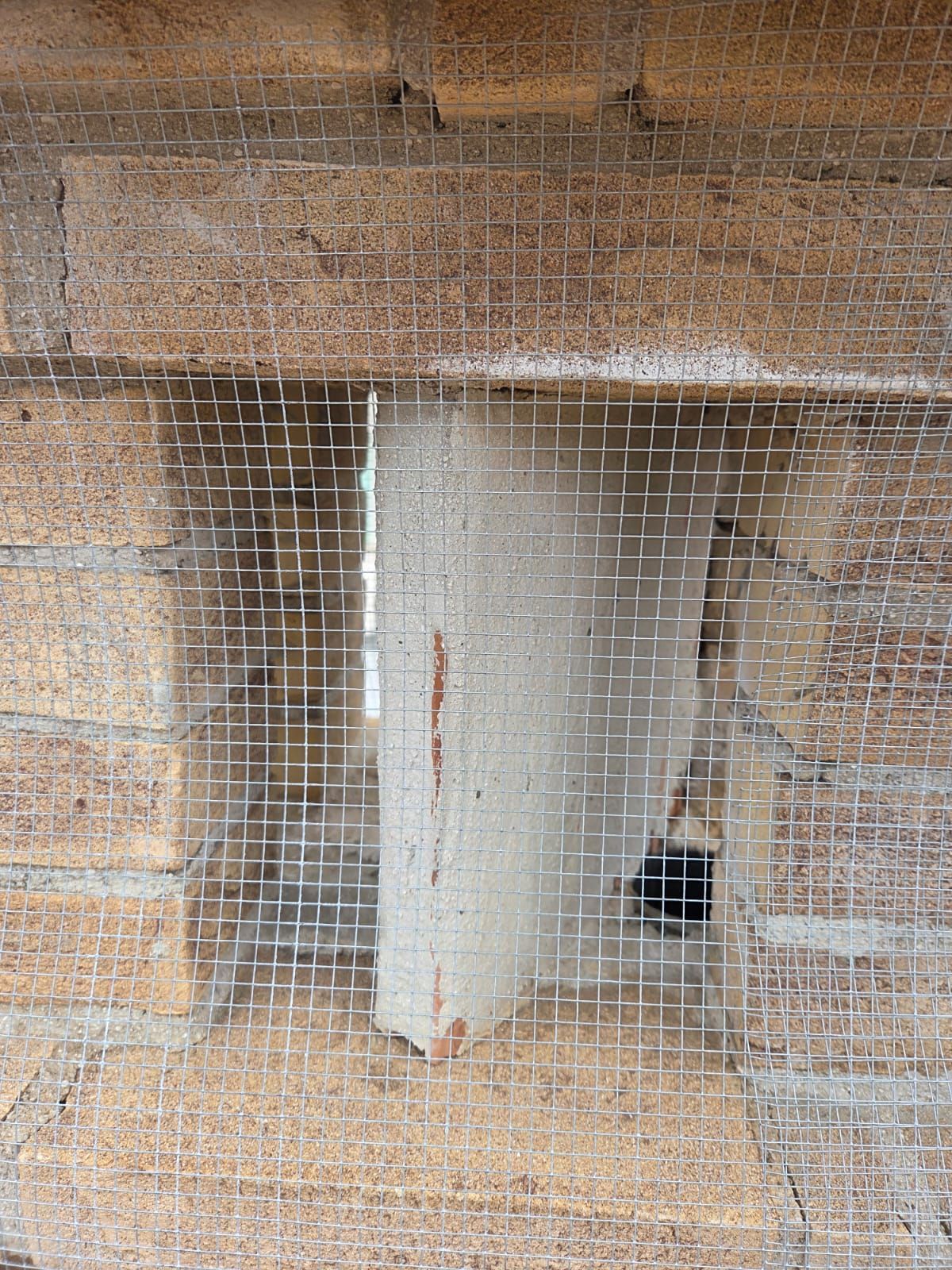 Muro in pietra con un'apertura rettangolare coperta da rete metallica, con una sezione di intonaco all'interno dell'apertura.