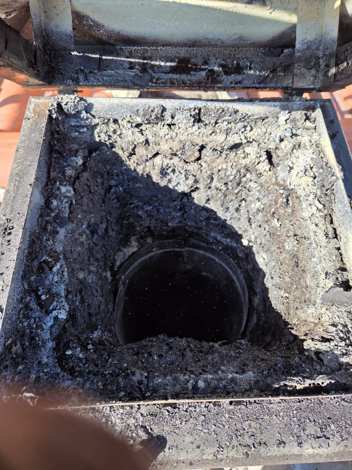 L'apertura di un camino è piena di fuliggine. Creosoto nero e cenere circondano un tubo di scarico scuro. La porta di accesso superiore è parzialmente aperta.