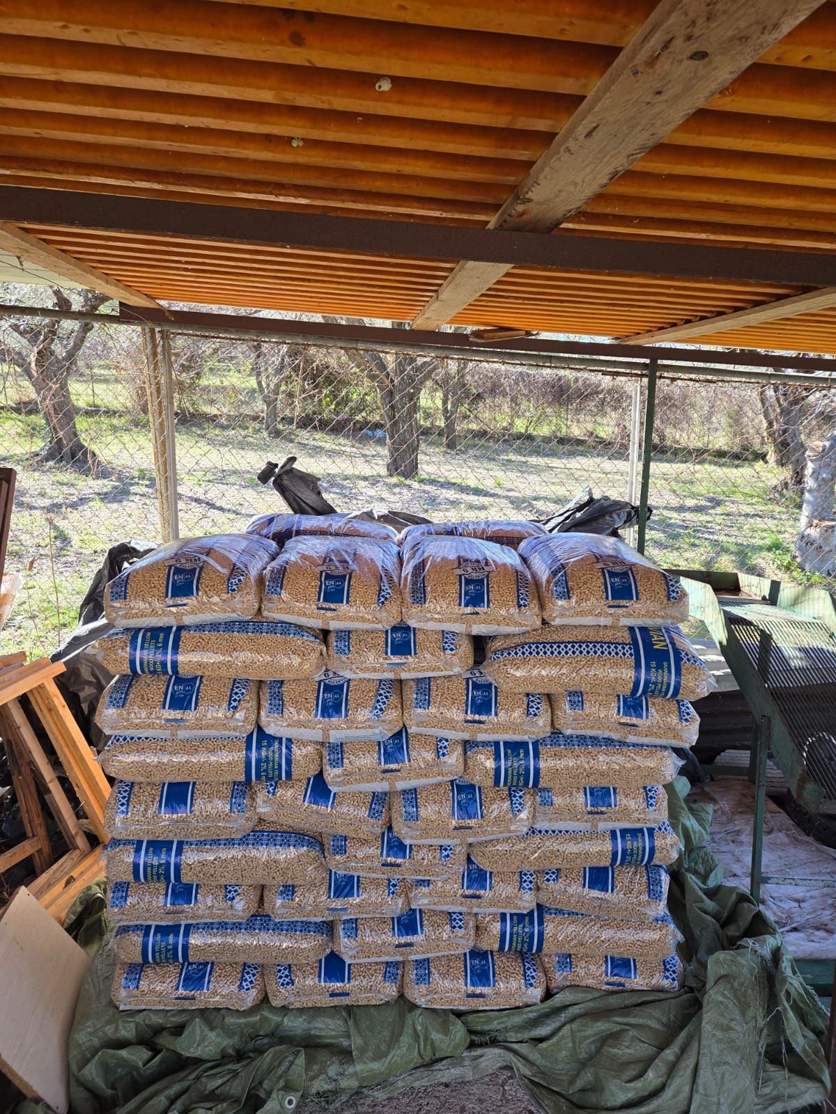 Pila di pellet di legno in sacchi sotto un riparo di legno, con uno sfondo esterno sfocato.