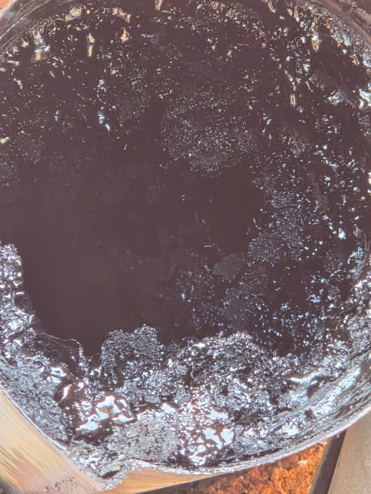 Liquido scuro e viscoso che riempie un contenitore metallico, con qualche bolla e solidificazione lungo i bordi.