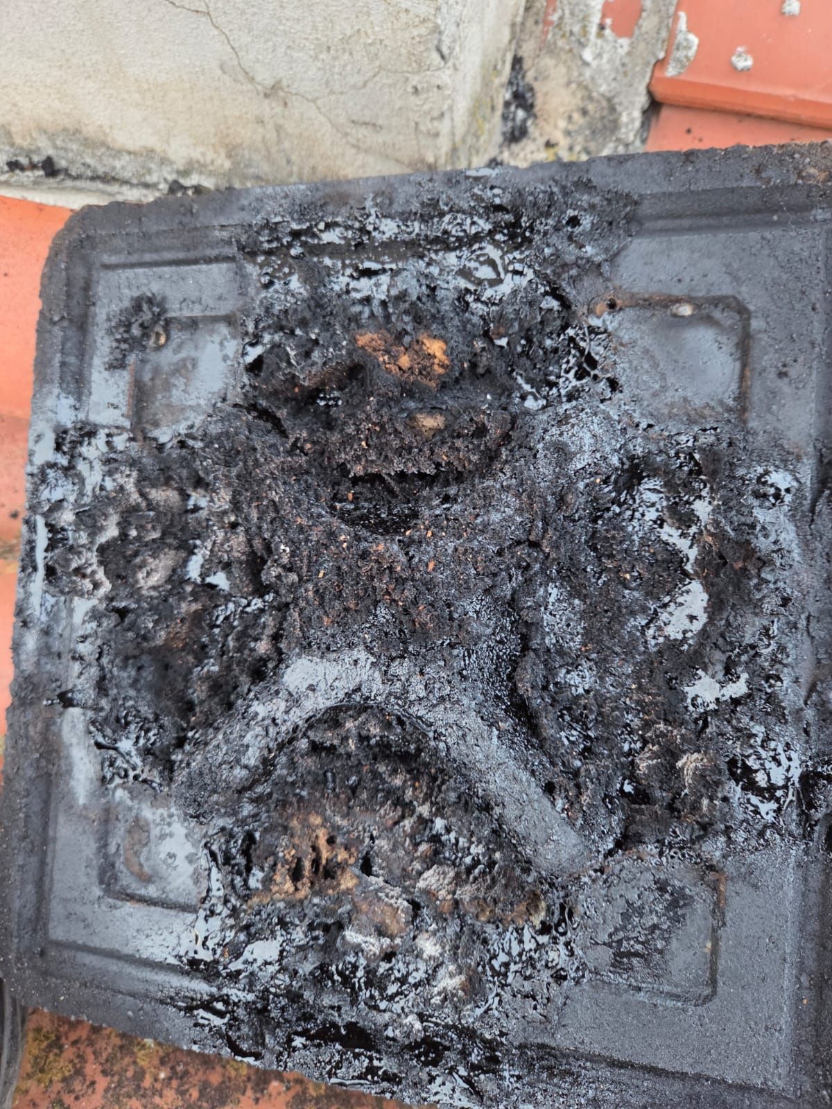 Contenitore di plastica gravemente ustionato su una superficie, con materiale carbonizzato e scuro e danni strutturali visibili.