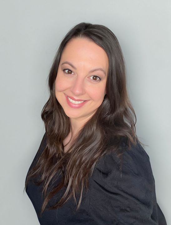 Woman with long brown hair smiles in front of a gray wall, wearing a black top.