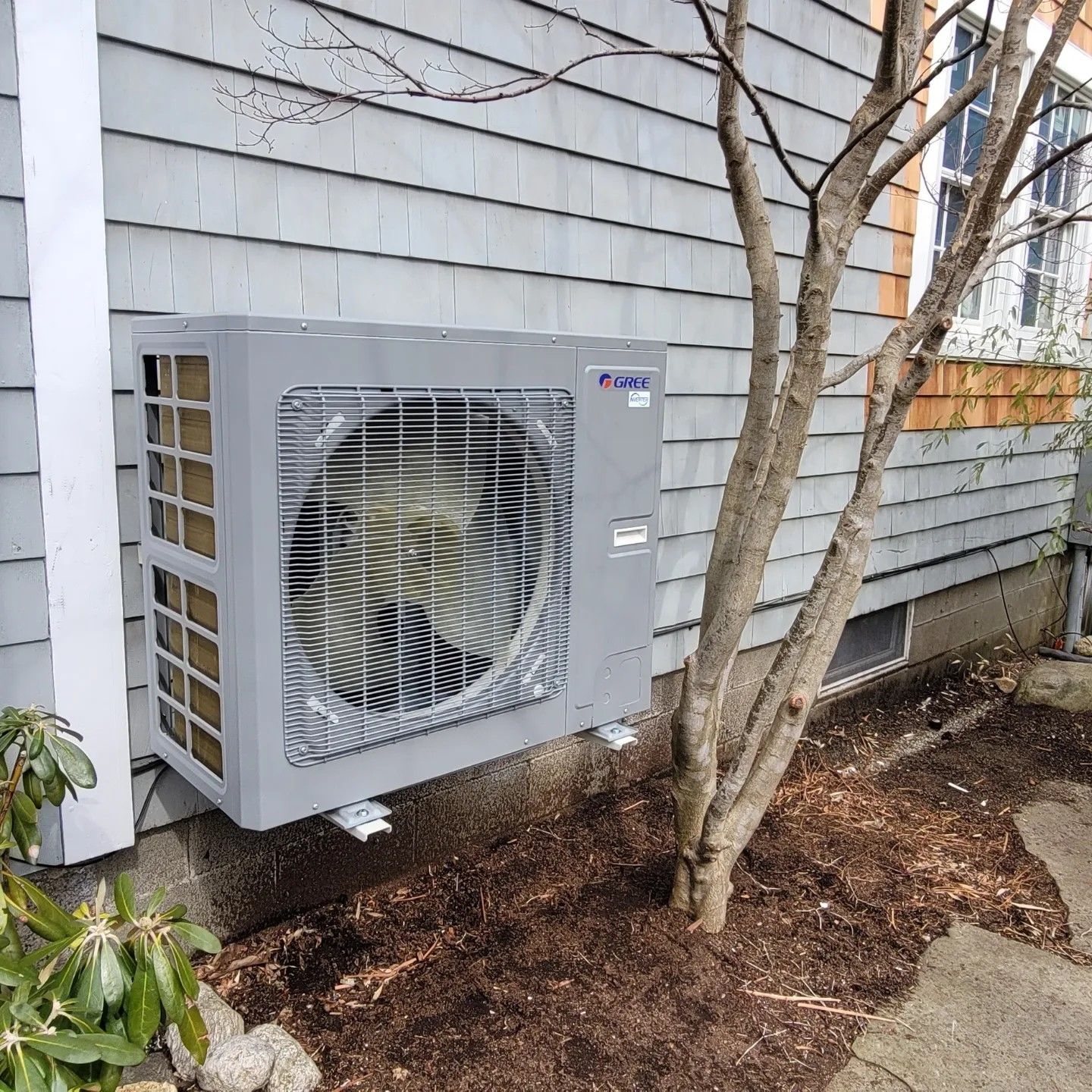 A large air conditioner is mounted on the side of a house next to a tree.