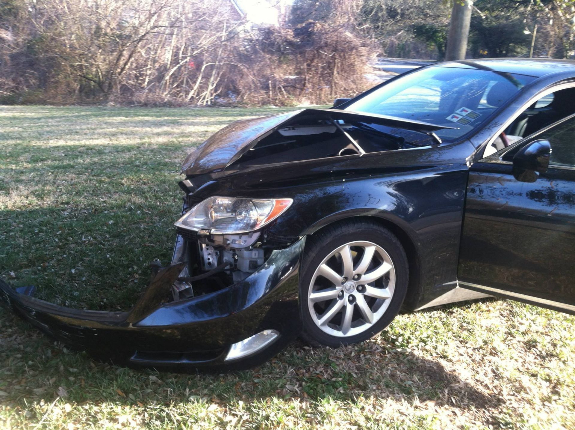 Black car with significant front-end damage, resting on grass next to a roadside area.