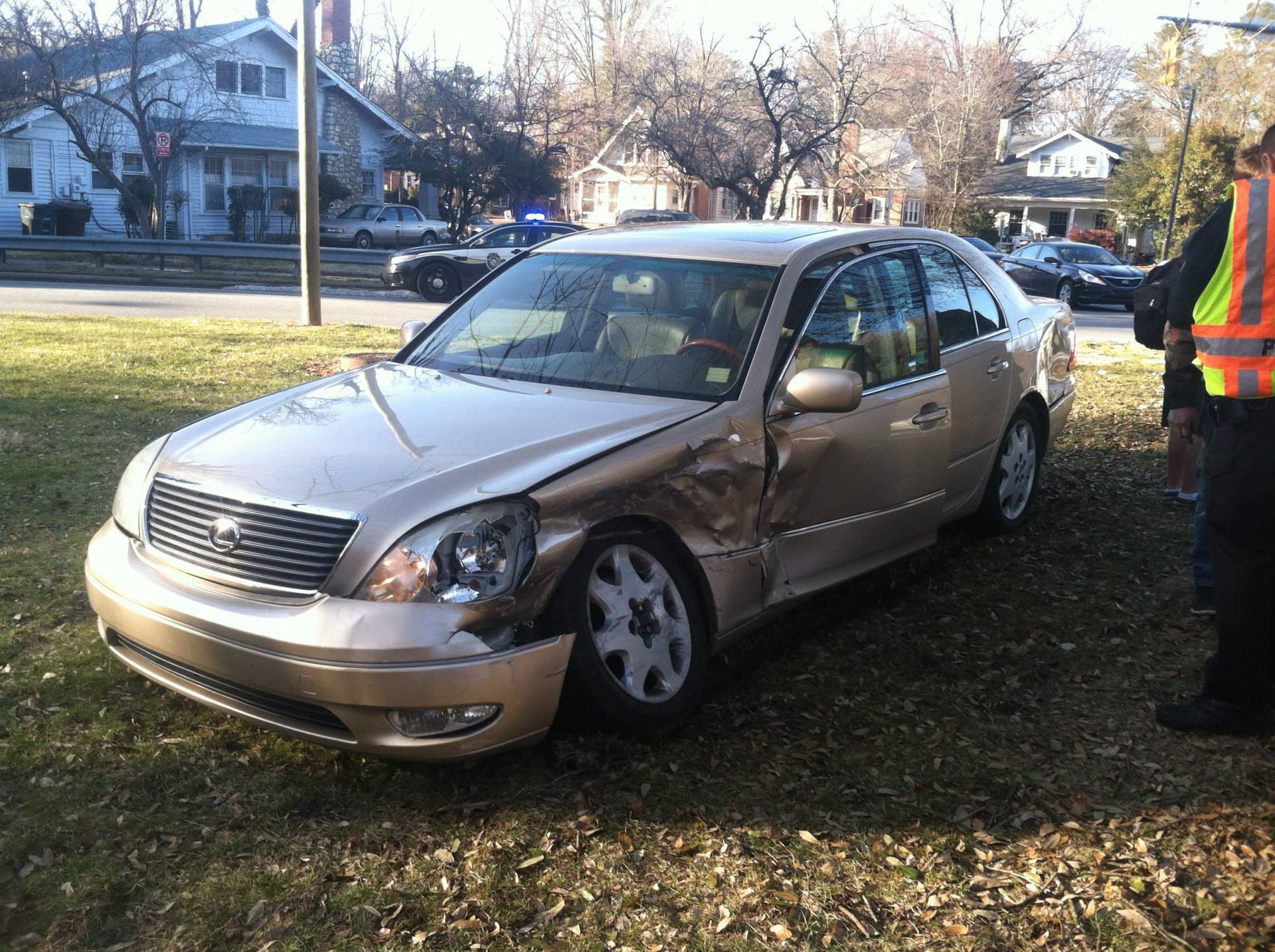 Gold sedan with front-end damage on a grassy area, possibly after a collision. A person in a safety vest stands nearby.