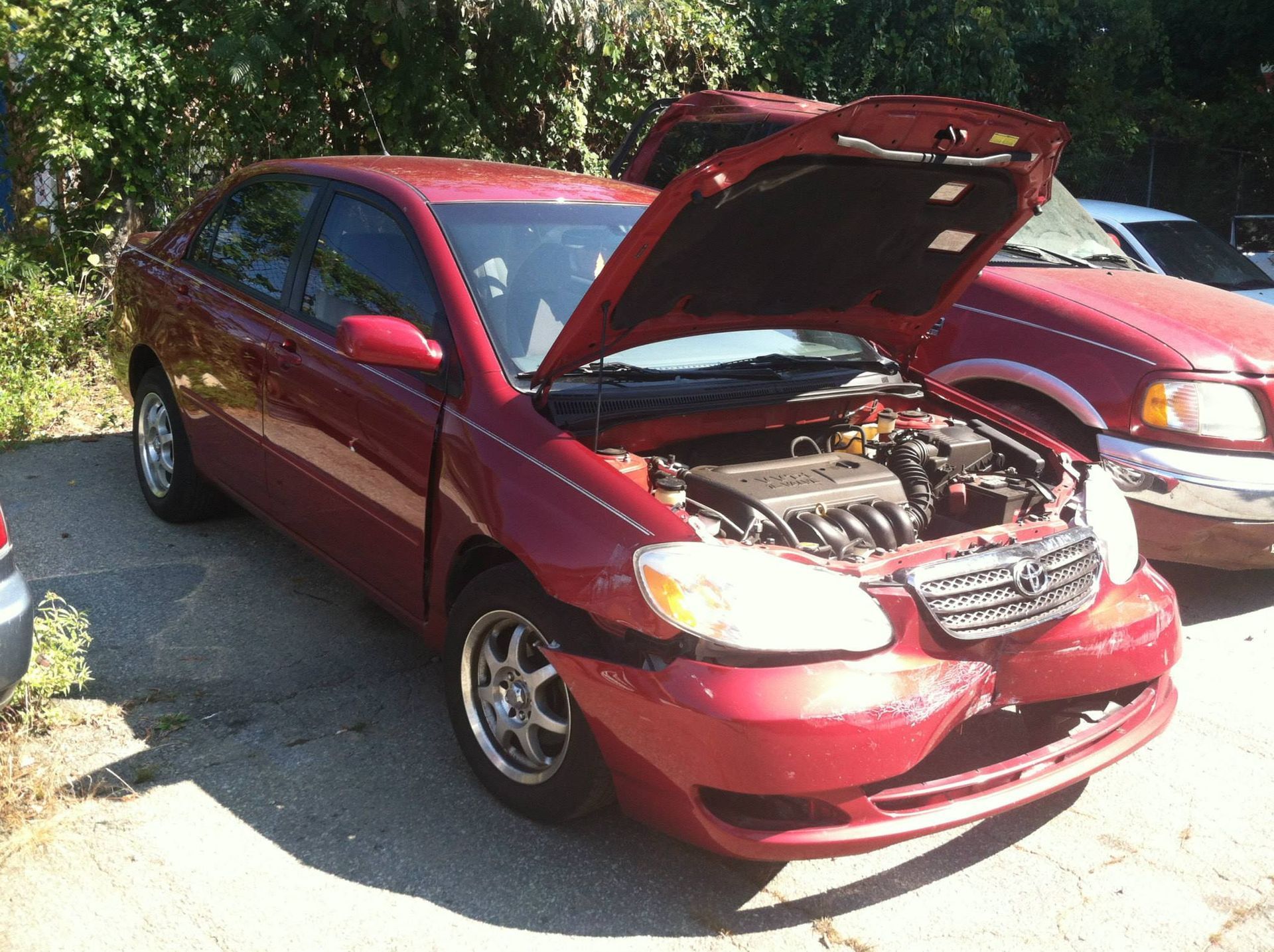 Red Toyota Corolla with front-end damage; hood open. Parked outdoors.