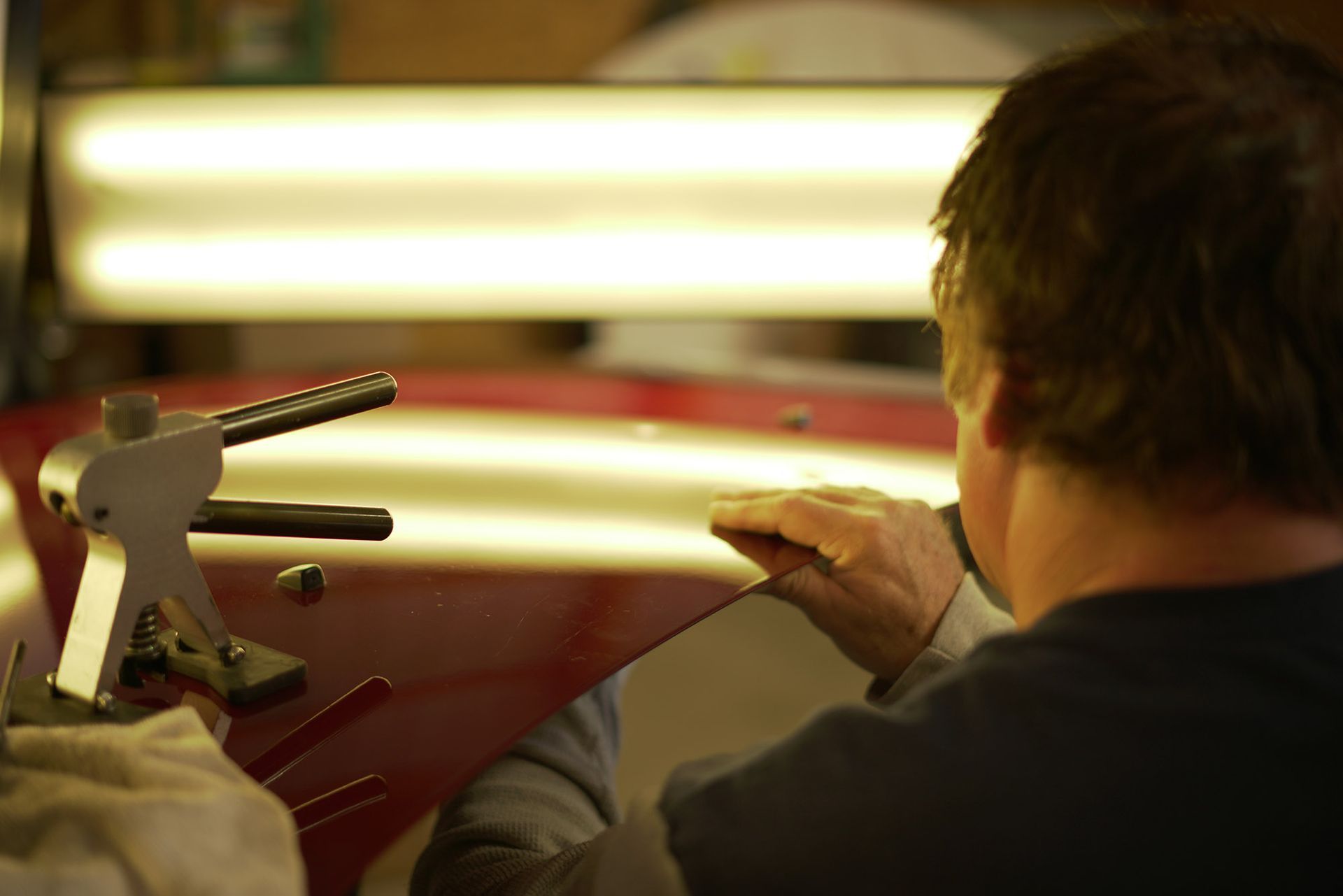 Person using a paintless dent removal tool on a red car panel under a bright light.