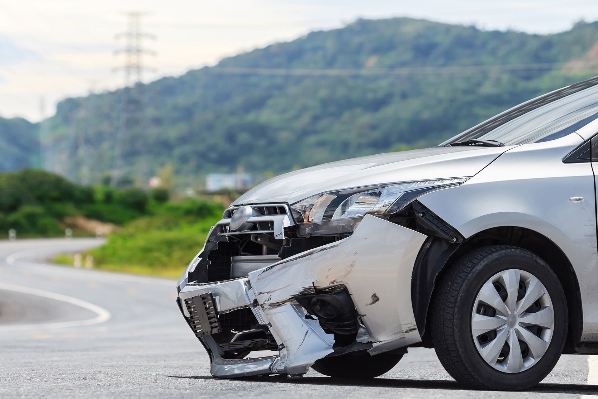 Silver car with significant front-end damage, roadside with green hillside in the background.