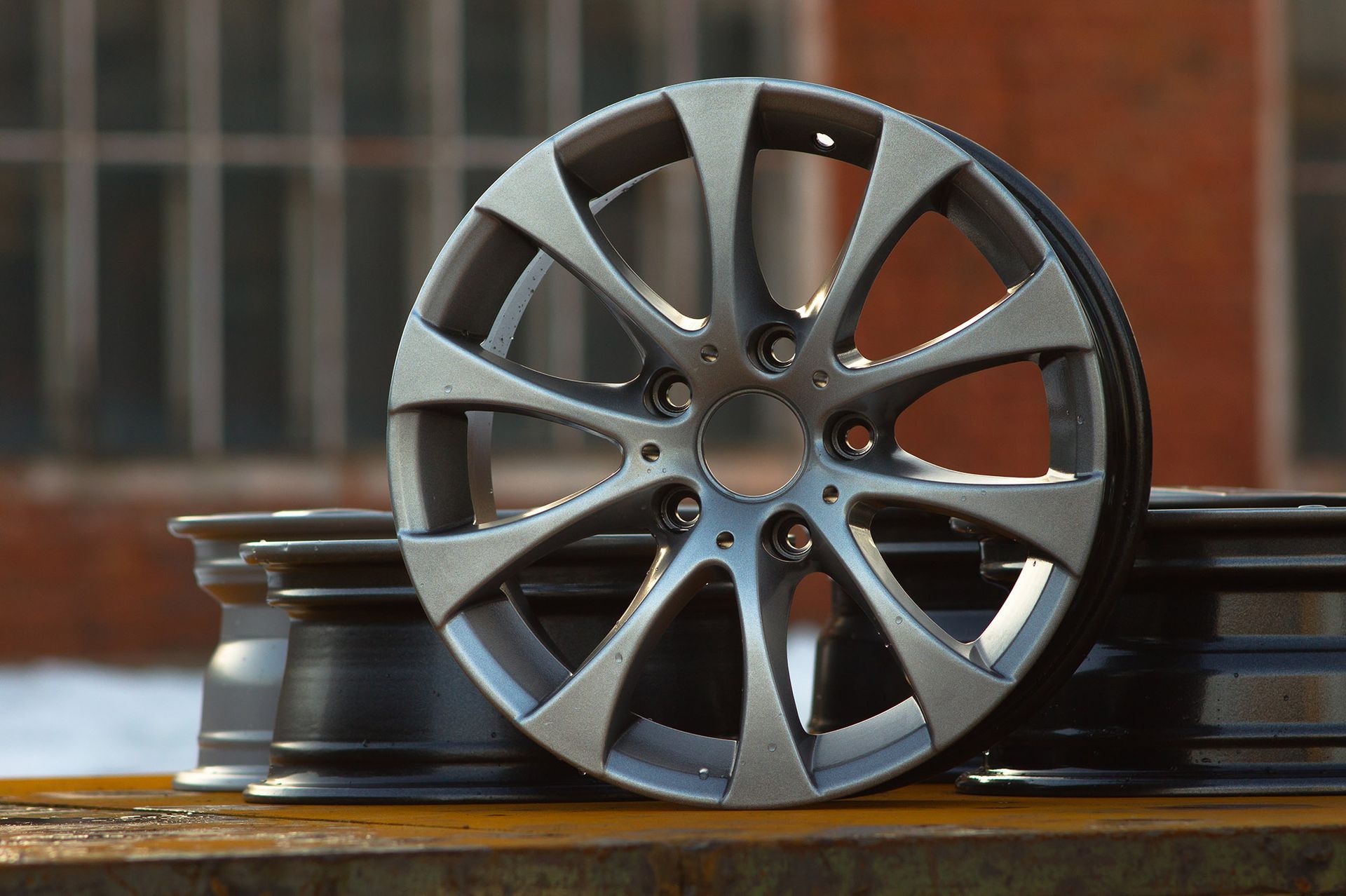 Silver alloy car wheel, close-up with a blurred background of a brick building. Other wheels in foreground.