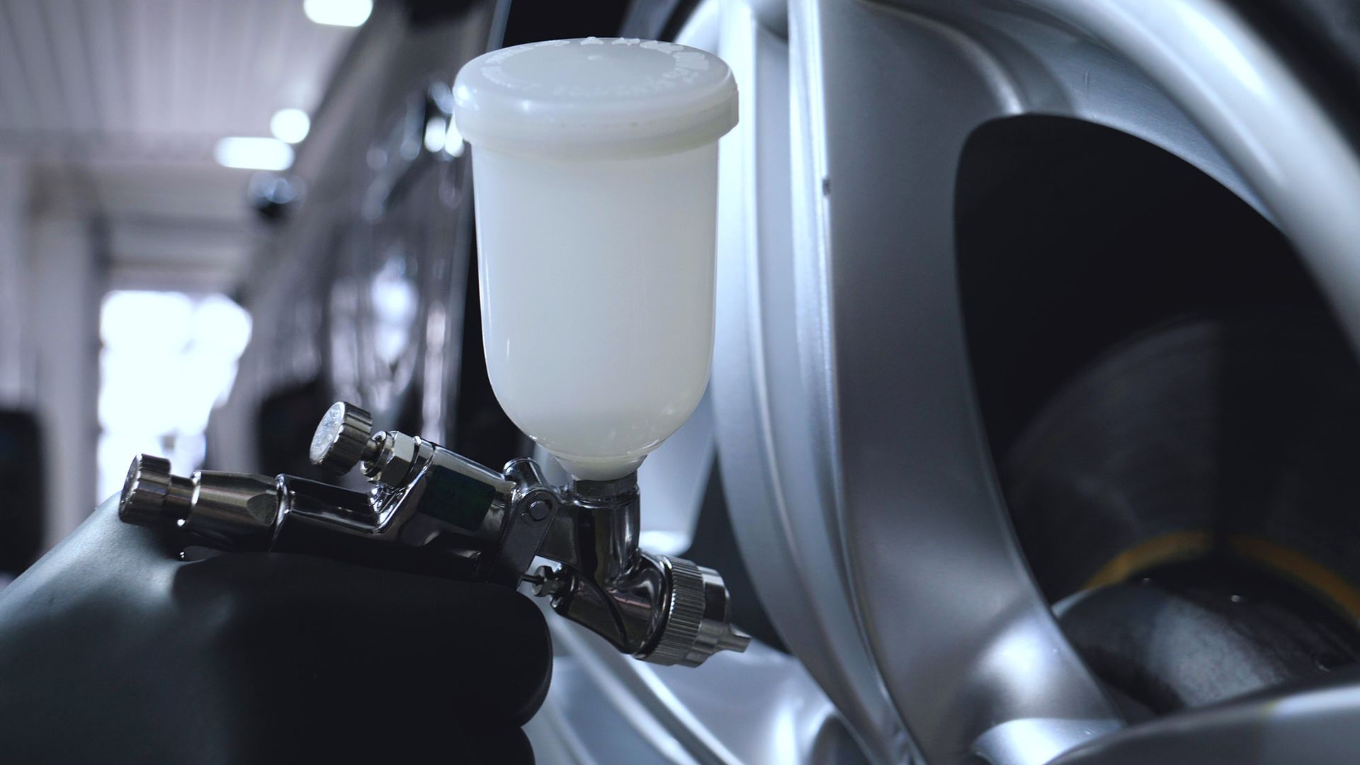 A person in a black glove spraying a car wheel with a paint gun in a workshop.