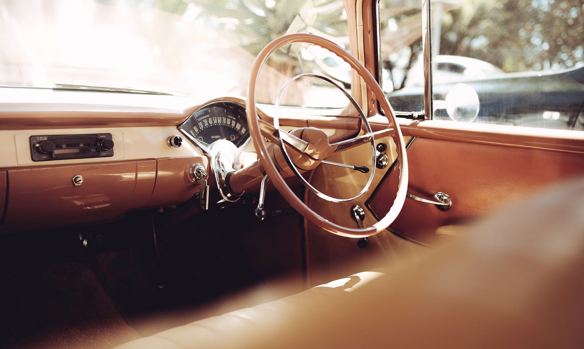 Interior of classic car: tan dashboard, steering wheel, and details, with a sunny, outdoor view.