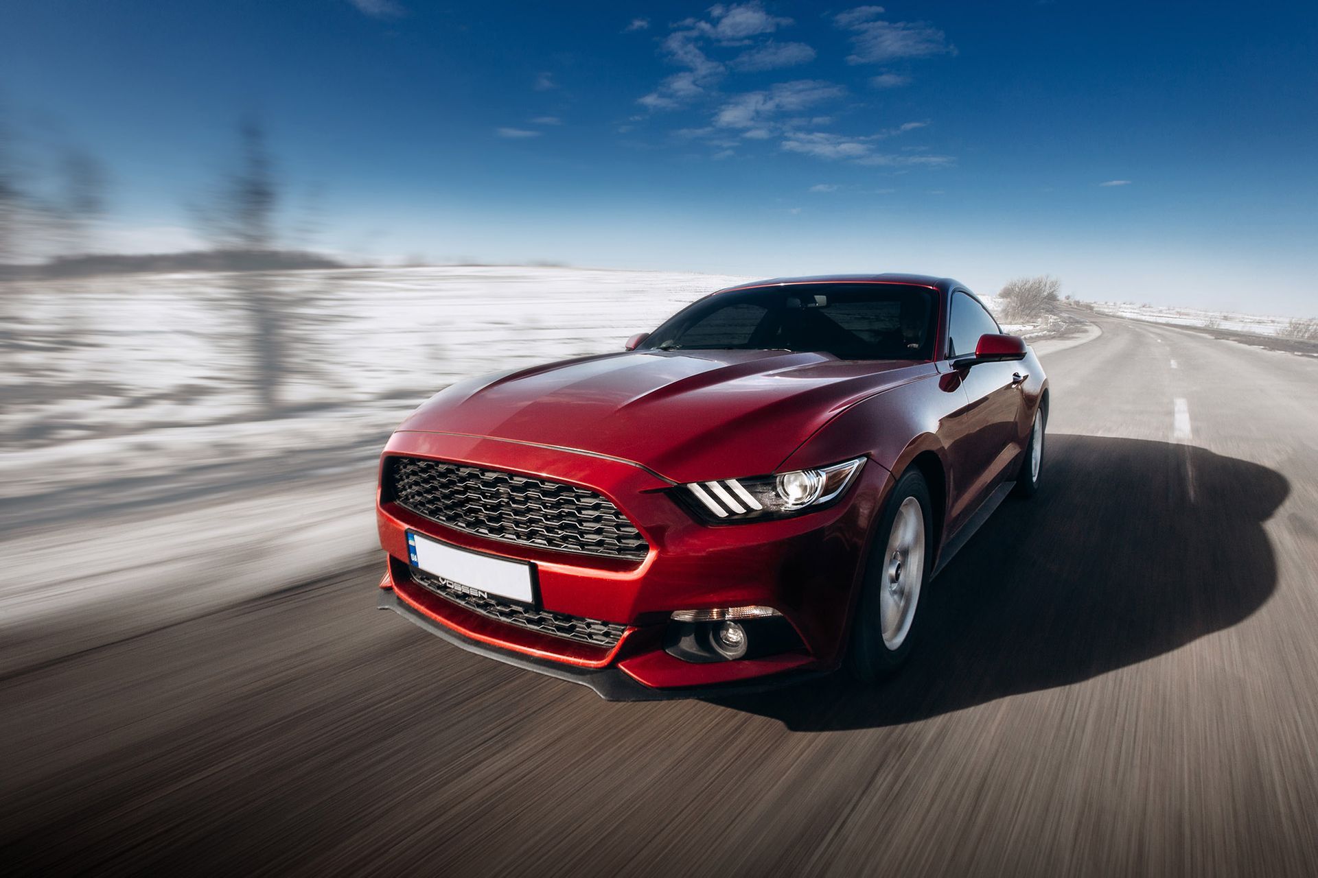 Red sports car driving on a road, snow-covered landscape, blue sky.