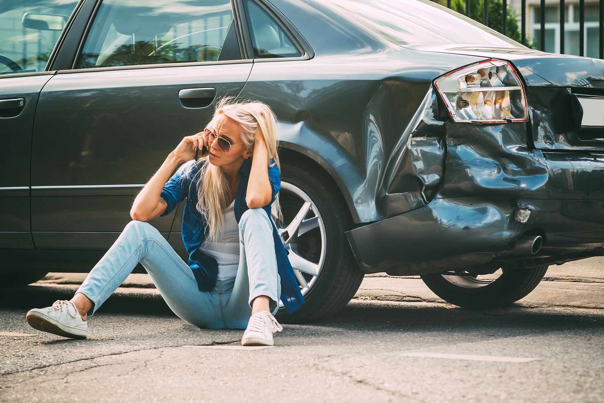 Woman sitting by damaged car, talking on phone, looking concerned.