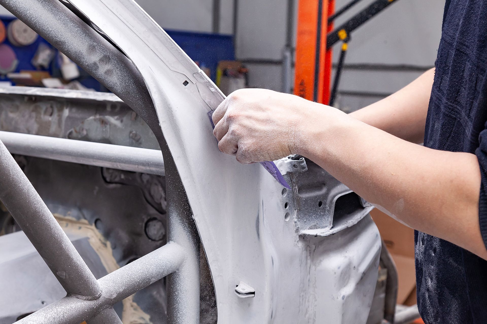 Person sanding a car body panel in a workshop, covered in gray dust.