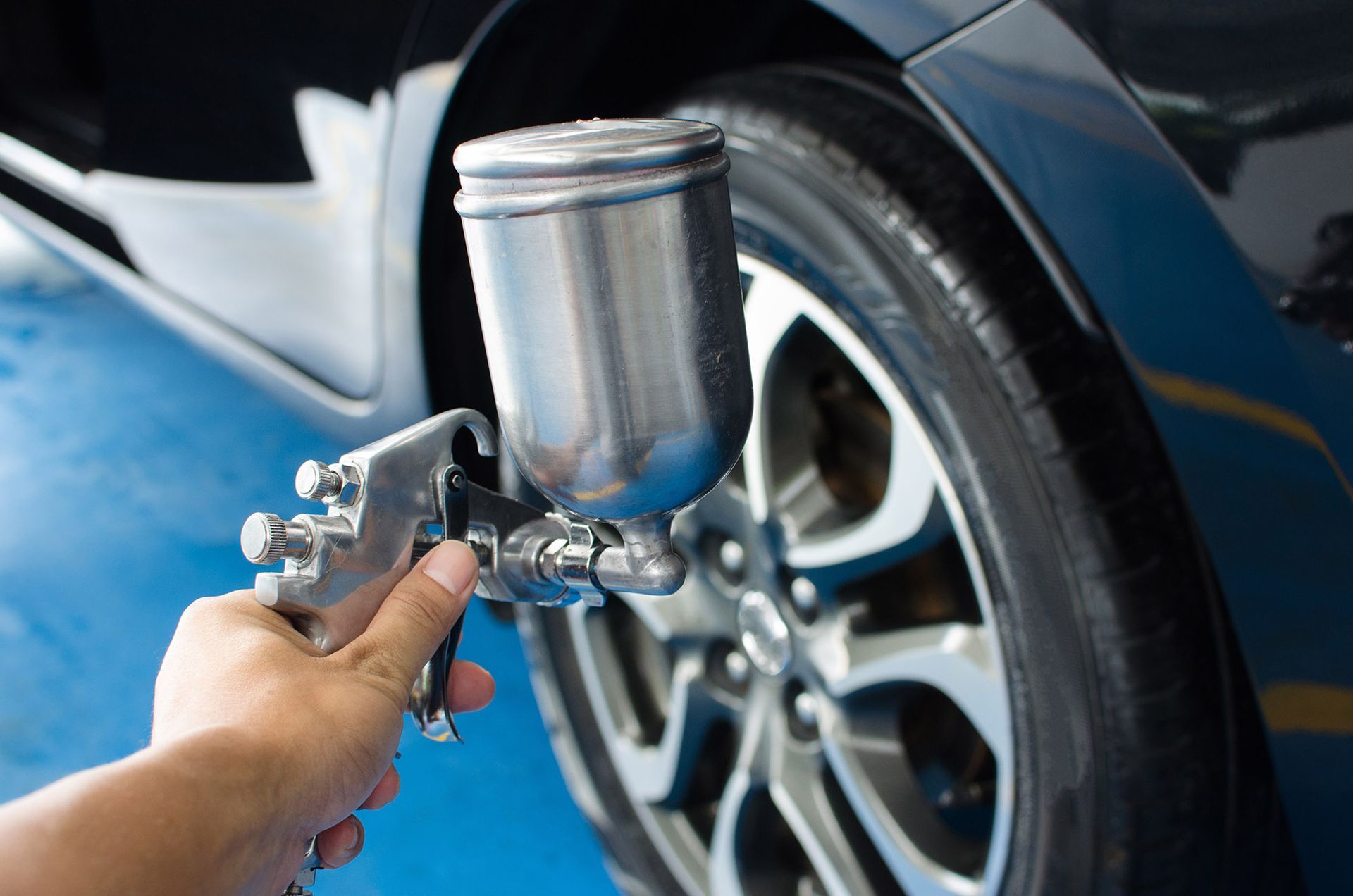 Hand holding a silver spray gun, painting a black car's tire in a blue-floored garage.